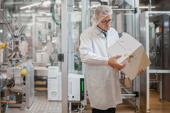 一名身着白色工服的工作人员正在检查纸箱的喷胶效果。背景中，配有Vision智能型热熔胶机的包装系统清晰可见。