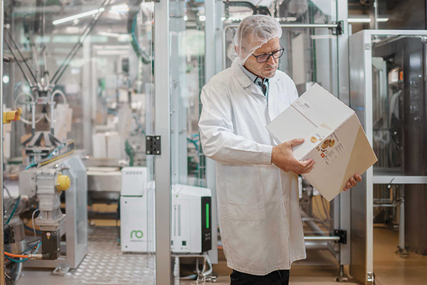 Man in een witte jas controleert de verlijming van een kartonnen verpakking. Op de achtergrond een verpakkingssysteem met de Vision-lijmsmeltmachine.