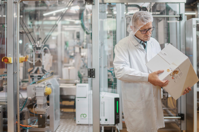 Expert vérifiant la qualité du scellage du processus de collage adhésif sur un emballage secondaire. À l'arrière-plan, un système d'emballage équipé du fondoir adhésif Vision.