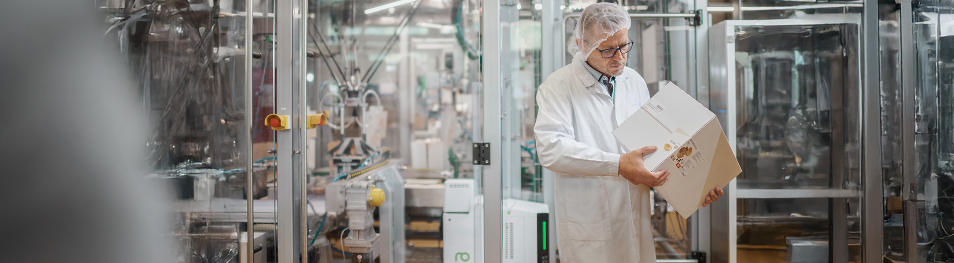 Expert vérifiant la qualité du scellage du processus de collage adhésif sur un emballage secondaire. À l'arrière-plan, un système d'emballage équipé du fondoir adhésif Vision.